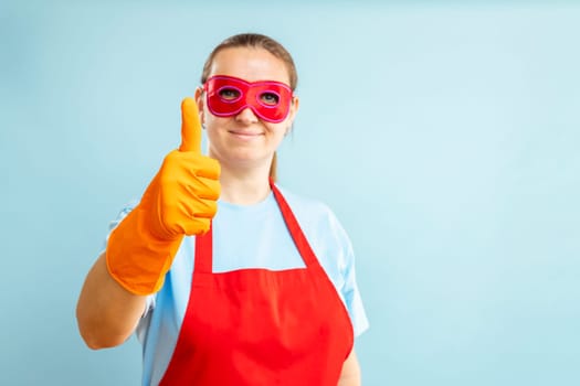 Young confident woman in red mask, gloves and apron showing ok sign with thumb up on blue background. Super housewife