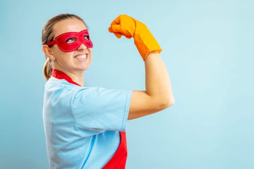 Confidence superhero house cleaner showing her powerful muscle arm with wearing red apron and eye mask on blue background