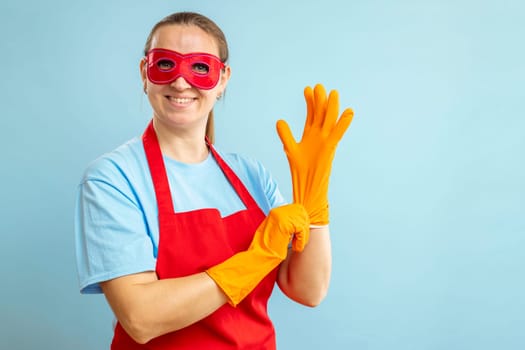 Young woman in red eye mask and apron puts on rubber gloves on blue background.