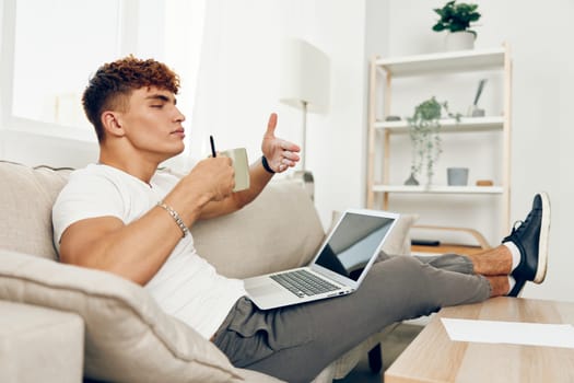 man interior curly computer looking adult caucasian chat t-shirt laptop cyberspace using online business white person phone cup typing couch