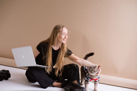 A business woman freelancer in a black is working, typing on a laptop apple and sitting on a white floor. blonde assistant in video production with computer MacBook and camera with cat