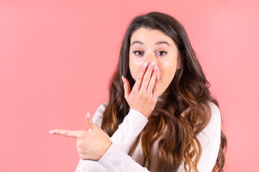 Brown-haired exited young woman closes face with palms pointing to shop. Lady with delighted expression shows boutique advertisement at pink wall