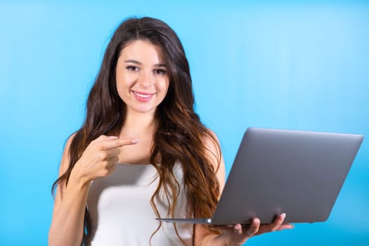 Brunette woman with amused and delighted expression looks at screen of laptop. Lady points to modern compact laptop with money win in blue studio
