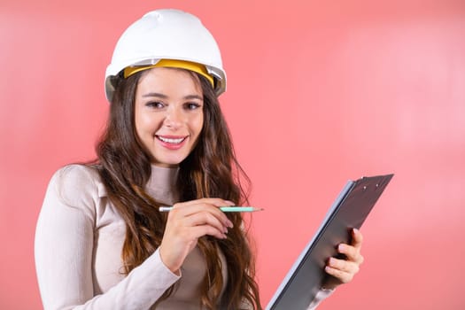 Brown-haired woman in white hardhat with smile prepares project for construction. Young lady engineer writes information on black clipboard