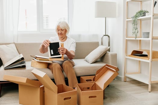 elderly woman sits on a sofa at home with boxes. collecting things with memories albums with photos and photo frames moving to a new place cleaning things and a happy smile. Lifestyle retirement. High quality photo