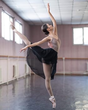 Graceful Asian ballerina in a beige bodysuit and black skirt is rehearsing in a dance class