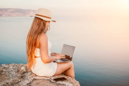 Digital nomad, Business woman working on laptop by the sea. Pretty lady typing on computer by the sea at sunset, makes a business transaction online from a distance. Freelance, remote work on vacation