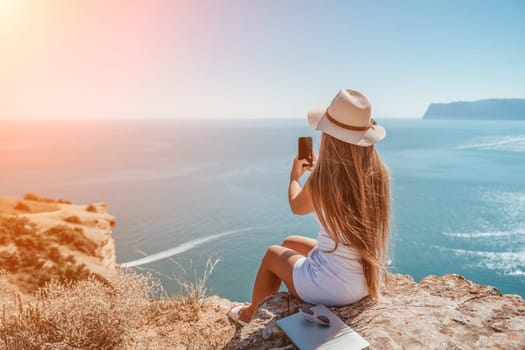 Successful business woman in yellow hat working on laptop by the sea. Pretty lady typing on computer at summer day outdoors. Freelance, travel and holidays concept.