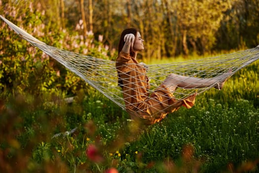 a happy woman in a long orange dress is resting sitting in a hammock at the dacha, smiling pleasantly looking away, illuminated by the summer sun during sunset. Horizontal photo. High quality photo