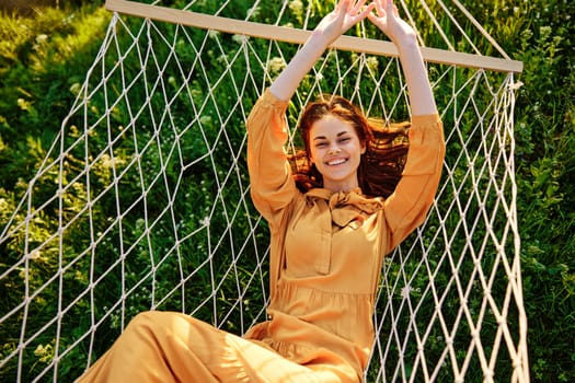 a beautiful, elegant woman lies in a long orange dress on a mesh hammock resting in nature, illuminated by the warm sunset light, joyfully raising her hands up. High quality photo
