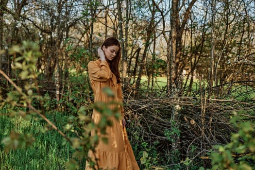 a beautiful, slender woman with long hair walks in the shade near the trees, dressed in a long orange dress, enjoying the weather and the weekend, straightening her hair with her hands. High quality photo