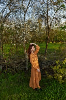 an elegant, sophisticated woman poses relaxed standing near a wicker fence at the dacha in a long orange dress touching her head with her hands. High quality photo