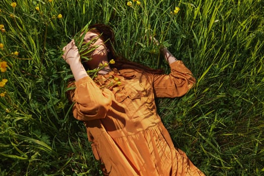 a calm woman with long red hair lies in a green field with yellow flowers, in an orange dress with her eyes closed, touching blades of grass with her hands, enjoying peace and recuperating. High quality photo