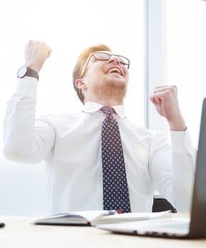 Happy young businessman raising hands in front of laptop