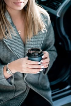 Happy woman coffee. she stands next to the car in the underground parking. Dressed in a gray coat, holding a glass of coffee in her hands, a black car