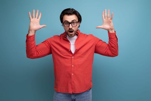 surprised young brunette man in glasses and a red shirt chatting on the phone raised his hands.