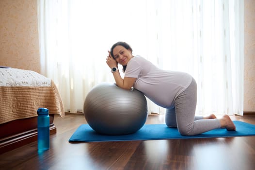 Happy adult pregnant woman leading active healthy lifestyle in pregnancy time, doing stretching exercises, standing on knees on a yoga mat leaning on a fitness ball at home, smiling looking at camera