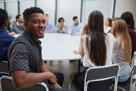 young African American man posing for the camera while sitting at a table against the background of his colleagues
