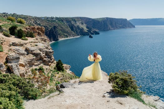 Woman in a yellow dress on the sea. Side view Young beautiful sensual woman in yellow long dress posing on a rock high above the sea at sunset. Girl in nature against the blue sky.