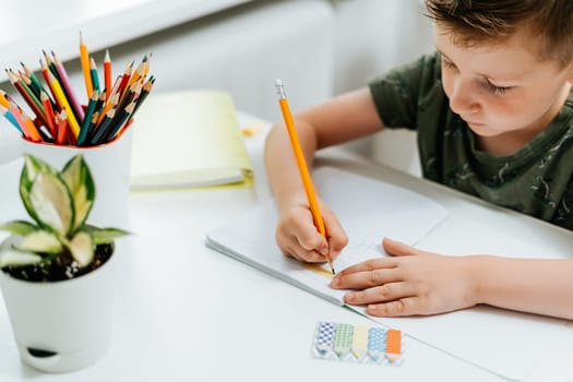 School kid boy studying at home with book, draw in notepad and doing homework. Thinking caucasian child siting at table with notebook. Back to school. Distance learning online education.