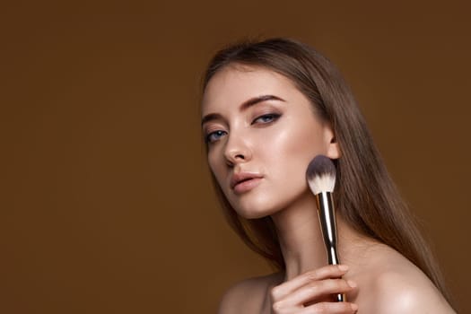 young woman applying blush on cheeks with make-up brush on beige background. copy space