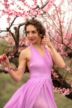 Woman peach blossom. Happy curly woman in pink dress walking in the garden of blossoming peach trees in spring.