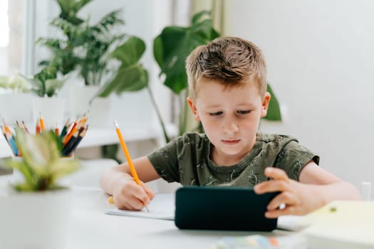 School kid boy studying at home with mobile smart phone and doing homework. Back to school. Thinking caucasian child with cellphone, pencil and training books. Distance learning online education.