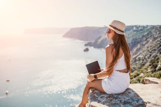 Successful business woman in yellow hat working on laptop by the sea. Pretty lady typing on computer at summer day outdoors. Freelance, travel and holidays concept.