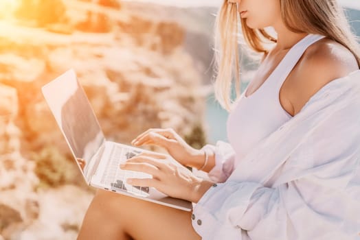 Digital nomad, Business woman working on laptop by the sea. Pretty lady typing on computer by the sea at sunset, makes a business transaction online from a distance. Freelance remote work on vacation