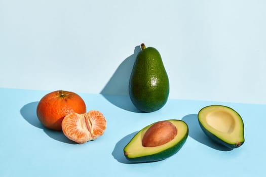 Composition of fresh fruits, mandarin, whole avocado and two halfs of cutted avocado on blue background. Mock up, two-colored pastel