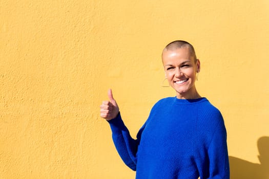 woman with a shaved head and wearing a blue sweater with her thumb up on a yellow wall in the background, concept of positive people, copy space for text