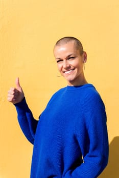 woman with a shaved head and wearing a blue sweater with her thumb up on a yellow wall in the background, concept of positive people, copy space for text