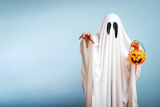 Halloween Concept. A white ghost with black eyes, made from a bedsheet with pumpkin basket for candy on blue background.