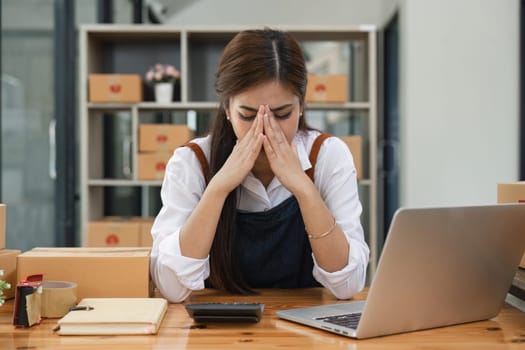 Small online business owner Asian woman wearing apron stressed headache taking notes with parcel box at home.