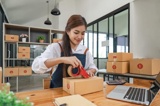 starting a small business Asian young woman business owner using masking tape to stick Close the parcel box, prepare for delivery through the shipping company according to the received online order.