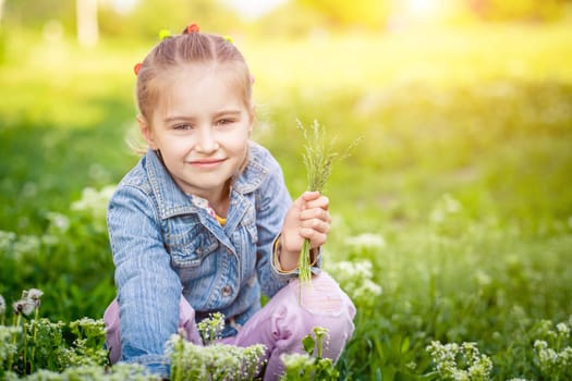 Cute small girl in jeans jacket is collecting bouquet of meadow flowers in green field