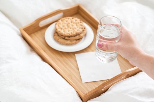 Female hand holding glass of water over tray with crackers on a bed