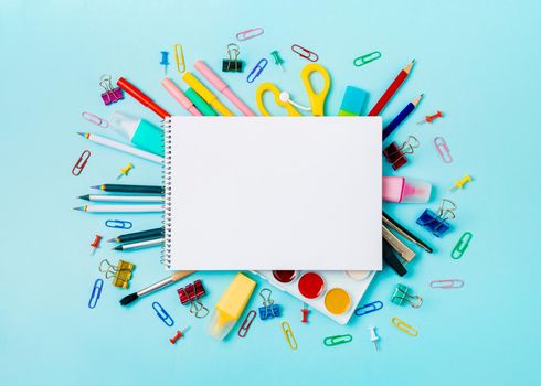 Notebook with stationary objects on blue background. Empty white paper sheet of notepad or sketchbook over shool supplies, isolated on blue. Copy space for text or design. Top view, flat lay