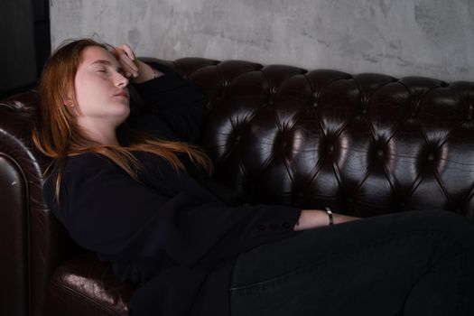 beautiful confident brunette woman in black on a brown leather couch in grey dark room. business woman. feminine. millennial people.