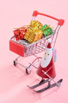 Christmas shopping concept, mini red shop cart trolley with Santa Claus toy and gift box isolated on pale pink background, blank copy space, close up