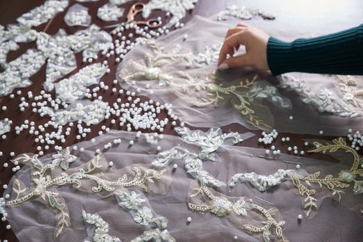 closeup of female hands of seamstress tailor sewing wedding dress using beads and lace in the studio