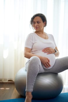 Vertical portrait of beautiful African American pregnant woman holding her belly, relaxing sitting on a fitball after prenatal stretching exercises at home. The concept of active and healthy lifestyle