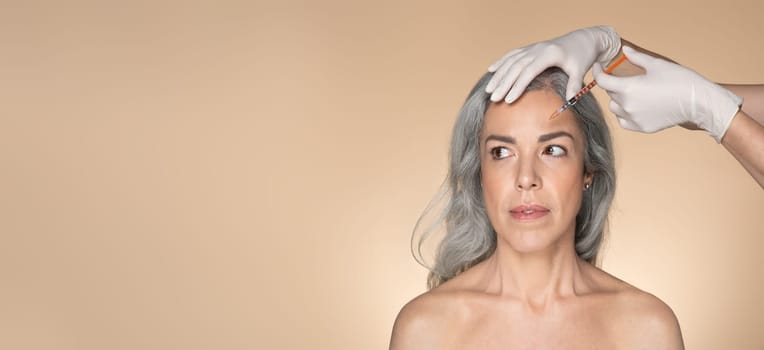 Senior caucasian woman on beauty procedure, cosmetologist hands in gloves making botox injection, beige background, panorama with free space. Anti-aging skin care, professional treatment