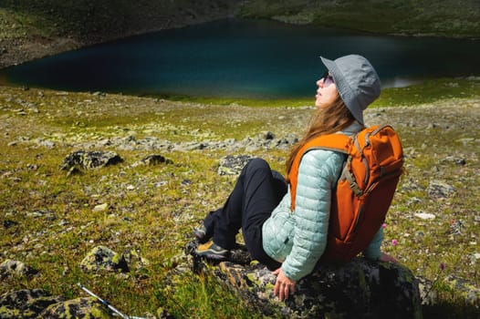 A female tourist sits and enjoys the view of the valley from the observation deck. The traveler has reached the top of the mountain and is relaxing with a backpack, against the backdrop of a mountain lake.