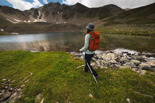 A young athletic woman walks through a green field with flowers next to a lake with picturesque mountains. A tourist with a backpack walks with trekking poles on a hike.
