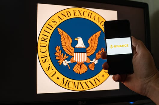 Kyiv, Ukraine - 06 June 2023: Man holds a smartphone with crypto exchange Binance on screen against the background of monitor with the SEC logo. S.E.C. sued on Binance through money laundering in BNB
