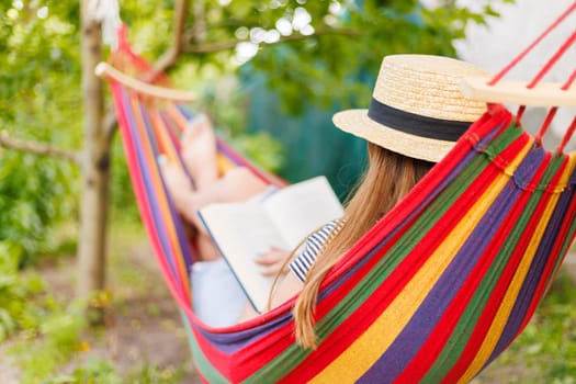 Young woman reading book while lying in comfortable hammock at green garden.