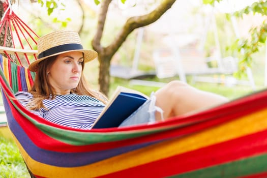 Young woman reading book while lying in comfortable hammock at green garden.