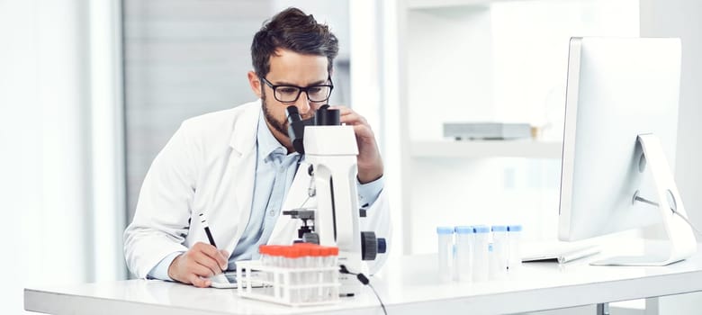 No distractions. a focused young male scientist making notes while looking trough a microscope and being seated inside a laboratory
