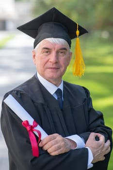 Portrait of an elderly man in a graduation gown and with a diploma in his hands outdoors. Vertical